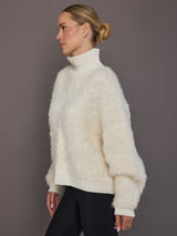 Model is wearing a cream oversized turtleneck sweater, side angle highlighting ribbed collar and drape.