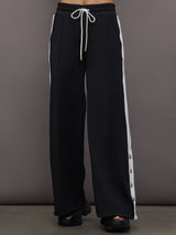 Model wears a black track pant with white stripe panel down sides and gold snaps running up to mid calf.
