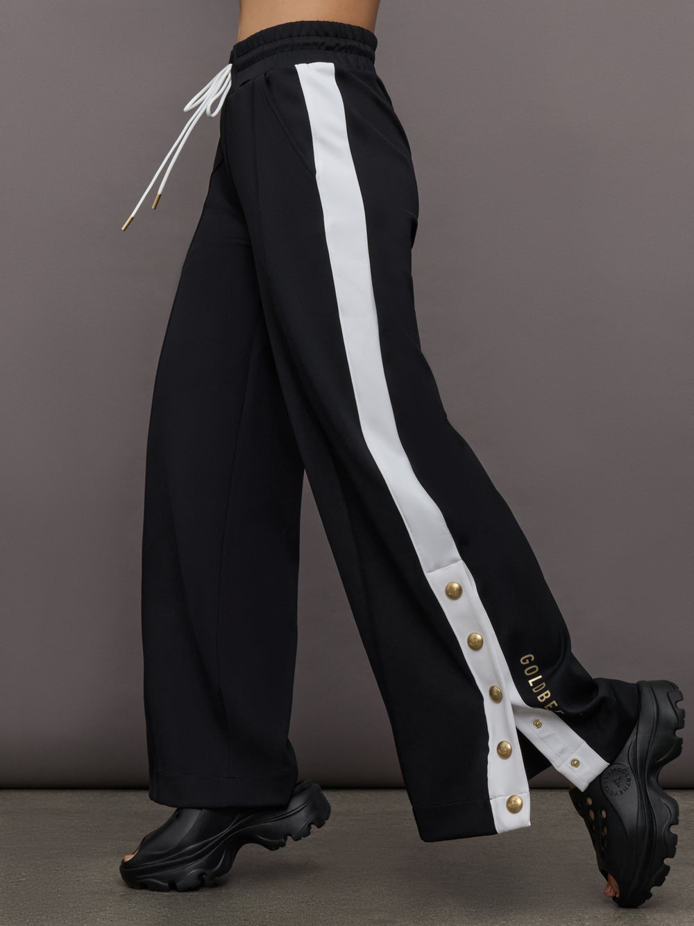 Model wears a black track pant with white stripe panel down sides and gold snaps running up to mid calf.