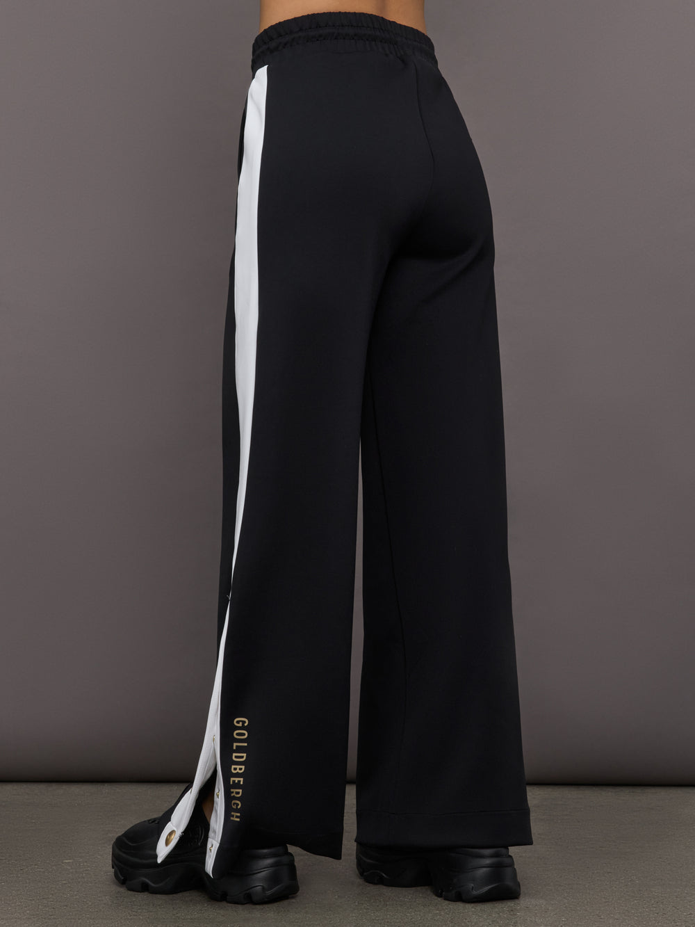 Model wears a black track pant with white stripe panel down sides and gold snaps running up to mid calf.