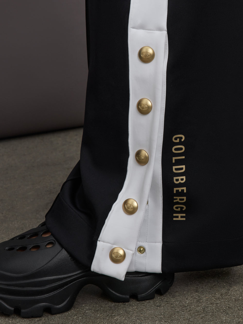 Model wears a black track pant with white stripe panel down sides and gold snaps running up to mid calf.