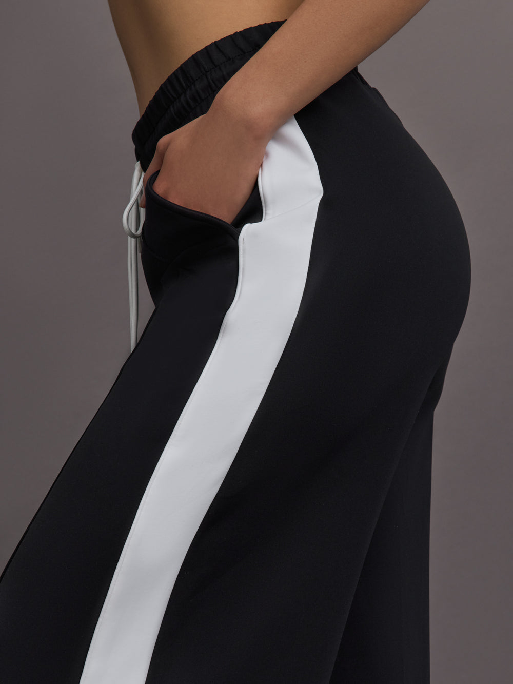Model wears a black track pant with white stripe panel down sides and gold snaps running up to mid calf.
