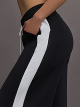 Model wears a black track pant with white stripe panel down sides and gold snaps running up to mid calf.