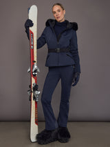 Full-body view of model wearing navy faux-fur ski jacket with matching pants and skis.