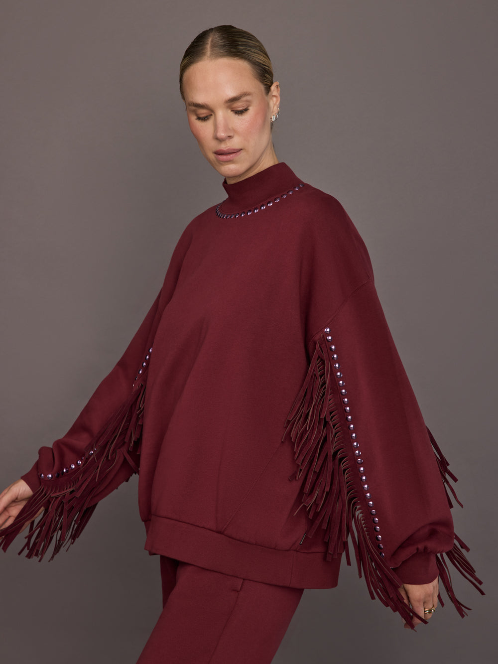 Model is wearing Frenzy Crew Neck Fringe Sweatshirt in Vino Rosso, side view showing sleeve fringe detail.