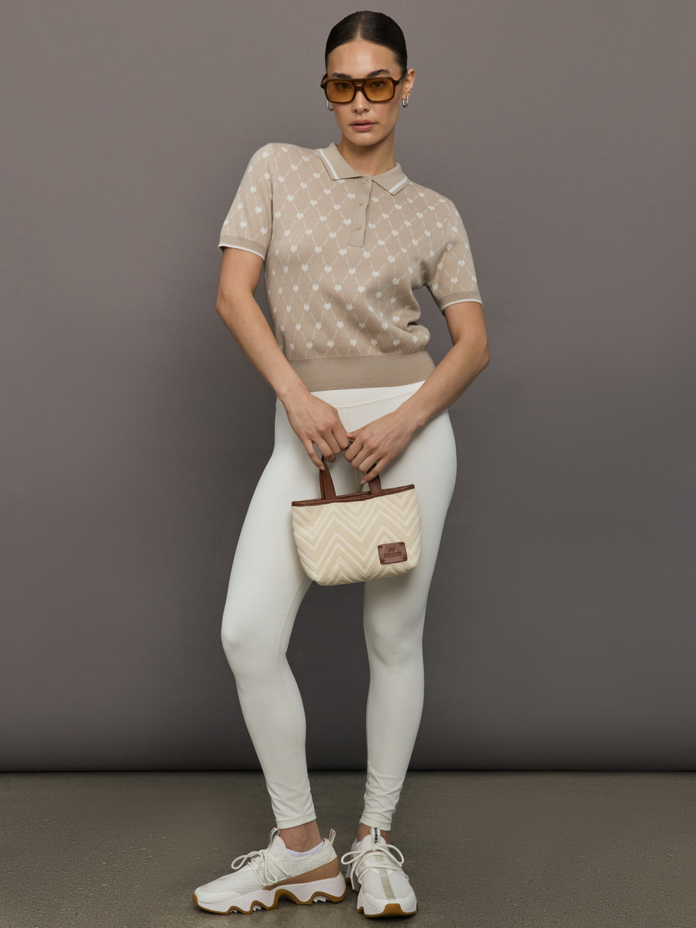 Woman wearing a beige polka dot top and white pants, holding a matching handbag against a gray background.