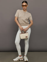 Woman wearing a beige polka dot top and white pants, holding a matching handbag against a gray background.