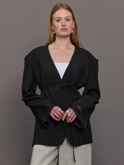 Model wears a wide shoulder black blazer with a deep v neck and drawstring closure. 