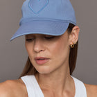 Model wears a denim colored baseball cap with an embroidered blue heart in front. 