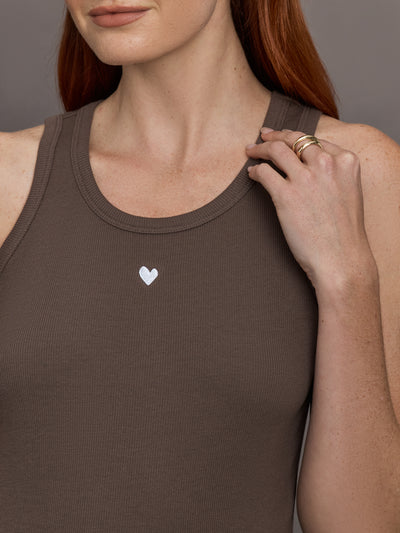 Model wears sleeveless tank top in a cocoa brown, featuring a scoop neckline and fitted silhouette and white heart in the upper center.