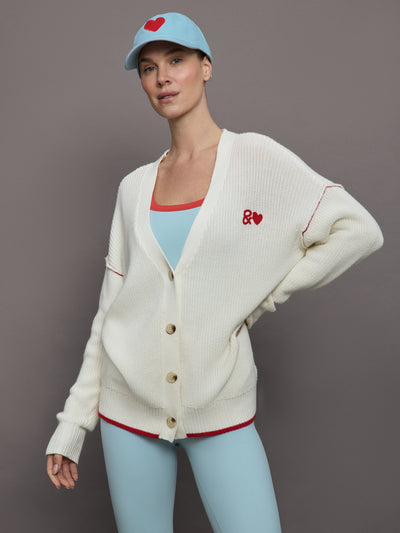 Model wears a deep V neck off white knit sweater with cuffed sleeves and red detailing on the left side of chest that reads "&" next to a heart.