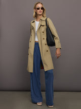 Woman wearing a beige trench coat and blue pants against a gray background