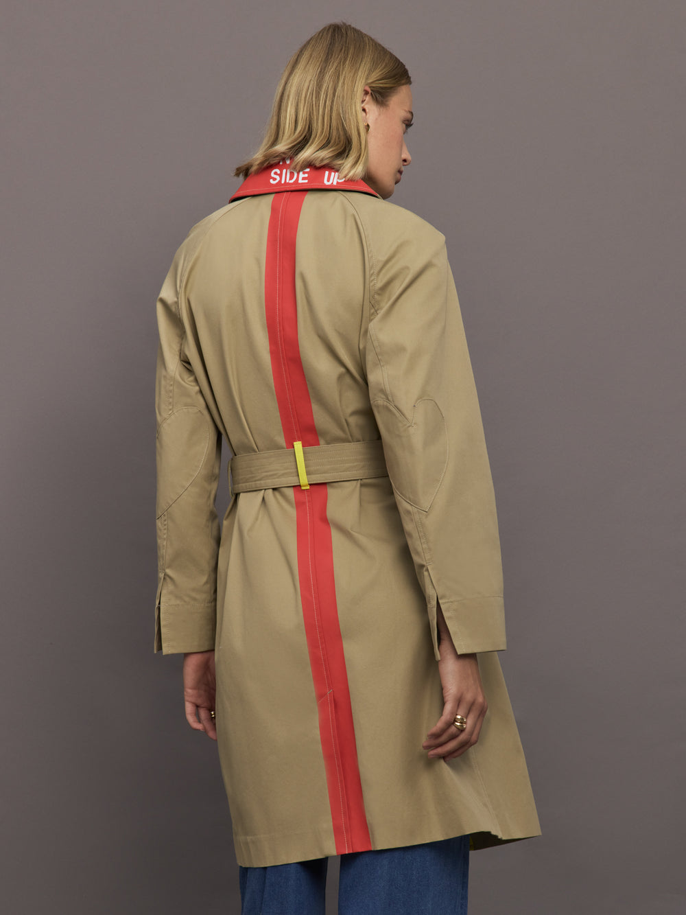 Person wearing a beige trench coat with red stripes and a visible brand logo on a gray background