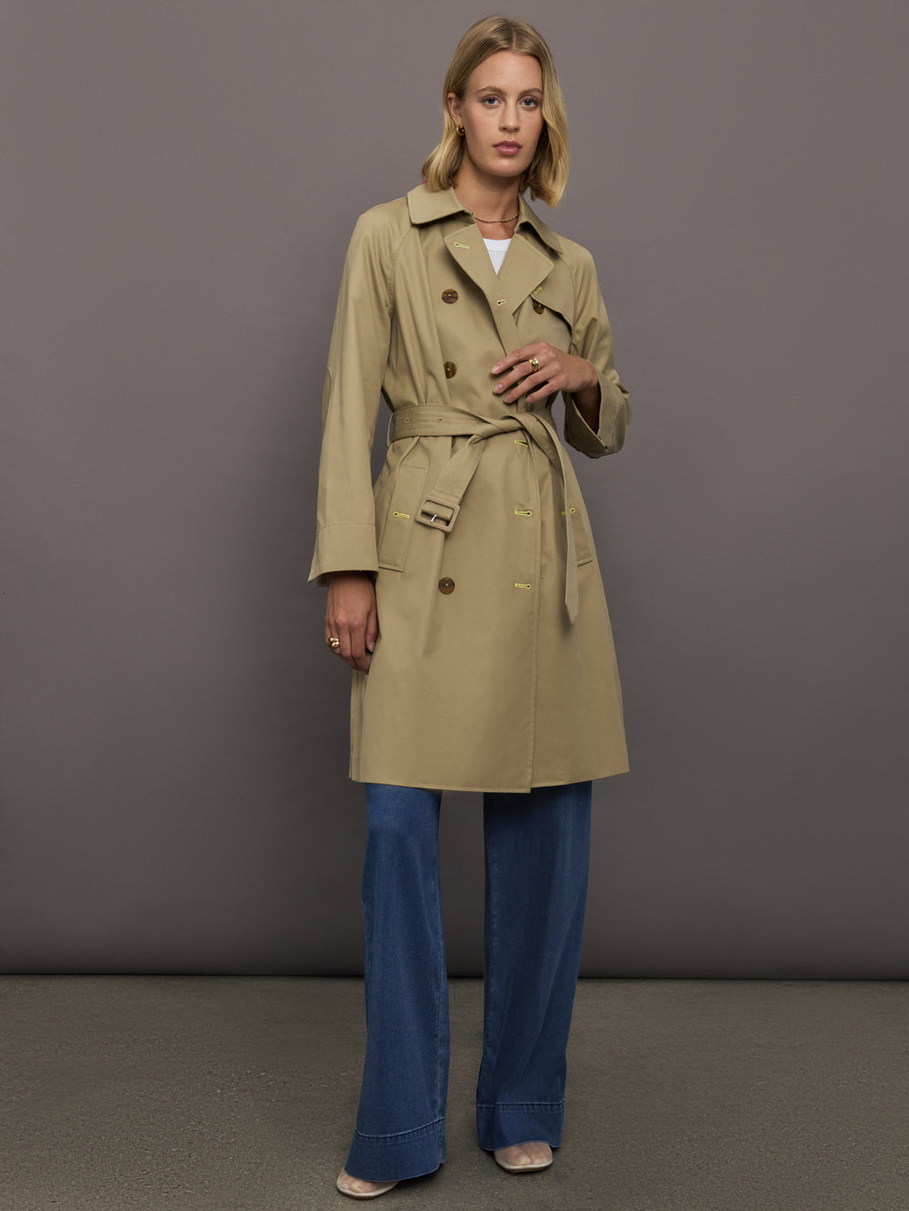 Woman wearing a beige trench coat and blue jeans against a gray background