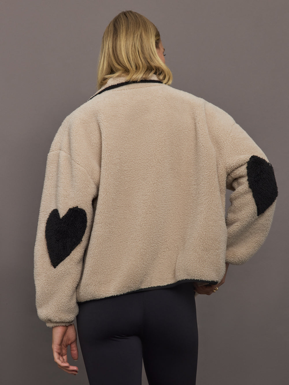 Model wears a camel colored fleece jacket with black embroidered heart on chest and elbows. 