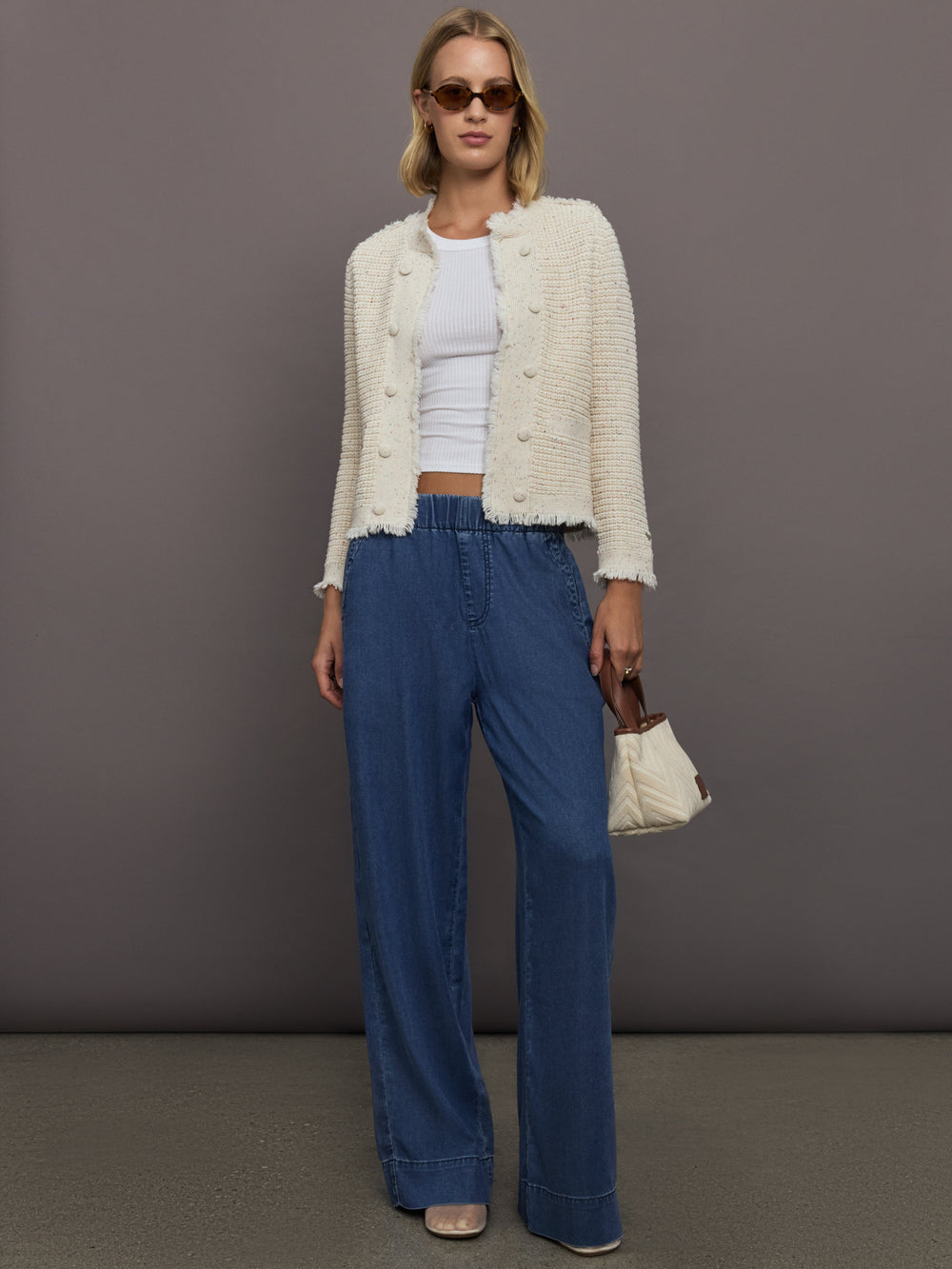 Woman wearing a white cardigan, white top, blue pants, and sunglasses against a gray background