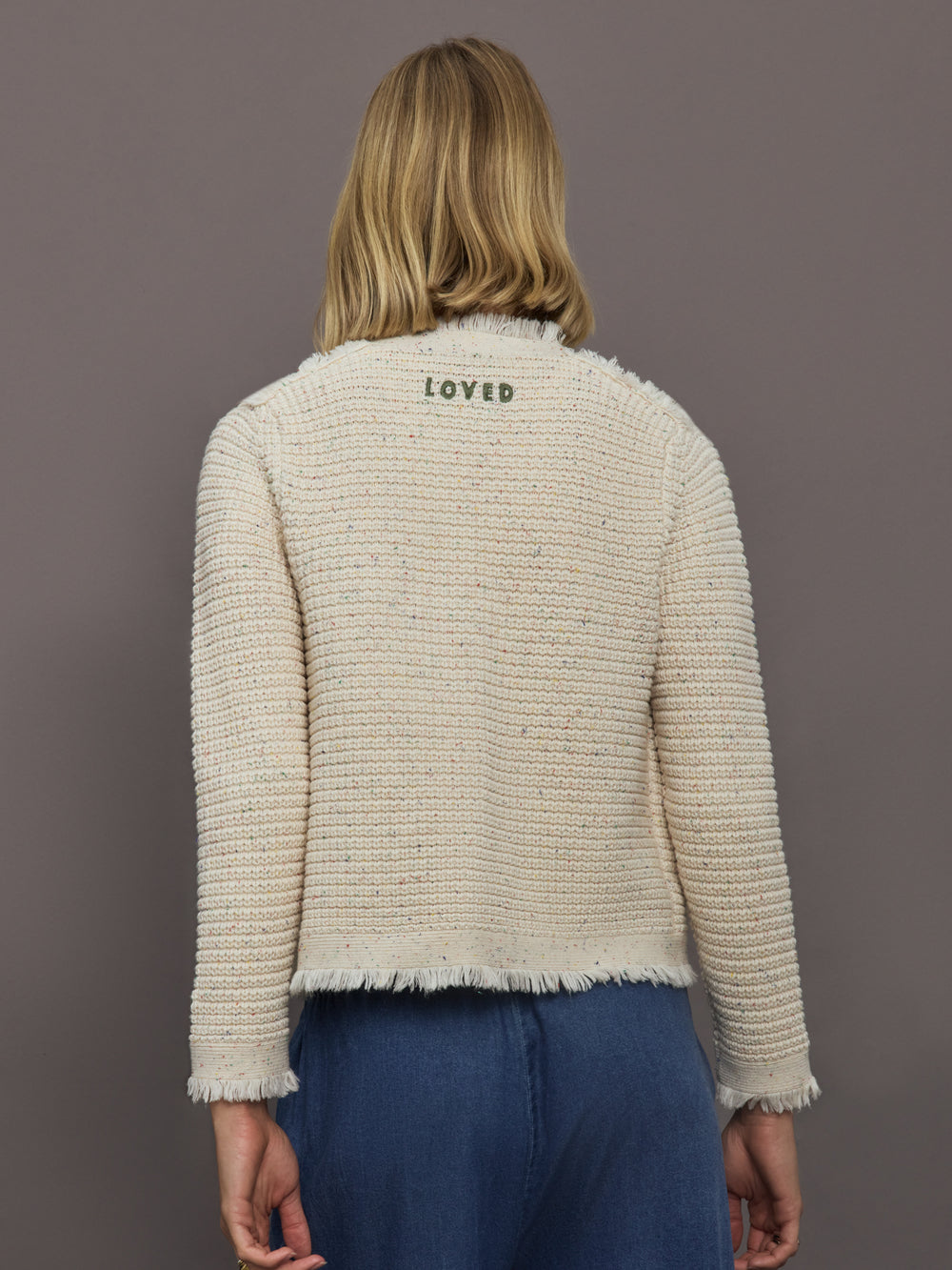 Person wearing a textured beige jacket with 'LOVED' embroidery on the back, standing against a plain background.