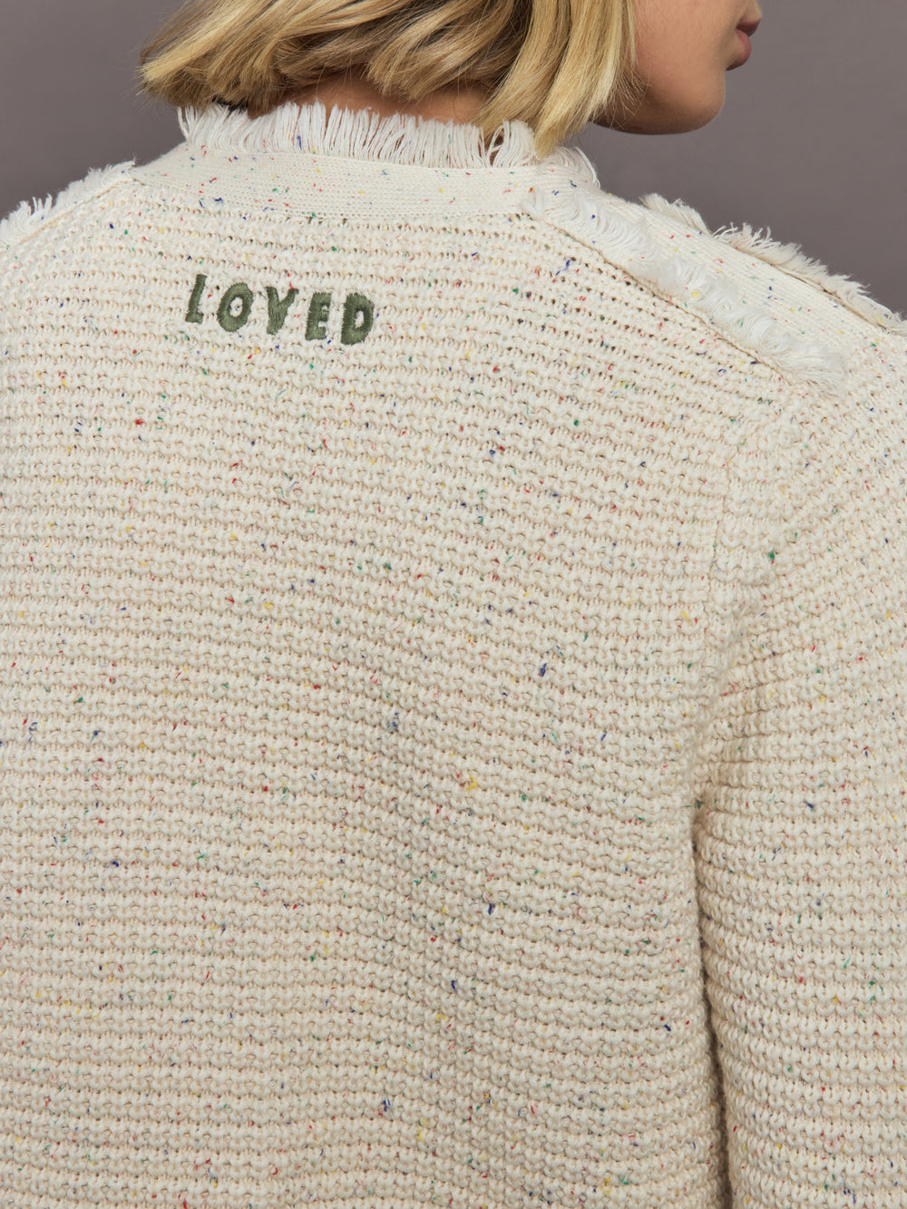 Back view of a textured beige sweater with 'LOVED' embroidered on it against a neutral background.
