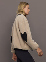 Model wears a camel colored fleece jacket with black embroidered heart on chest and elbows. 