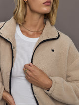 Model wears a camel colored fleece jacket with black embroidered heart on chest and elbows. 