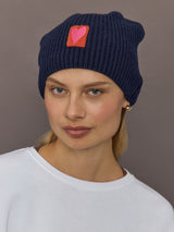 Model wearing indigo blue cotton-cashmere beanie with pink heart patch, front view.