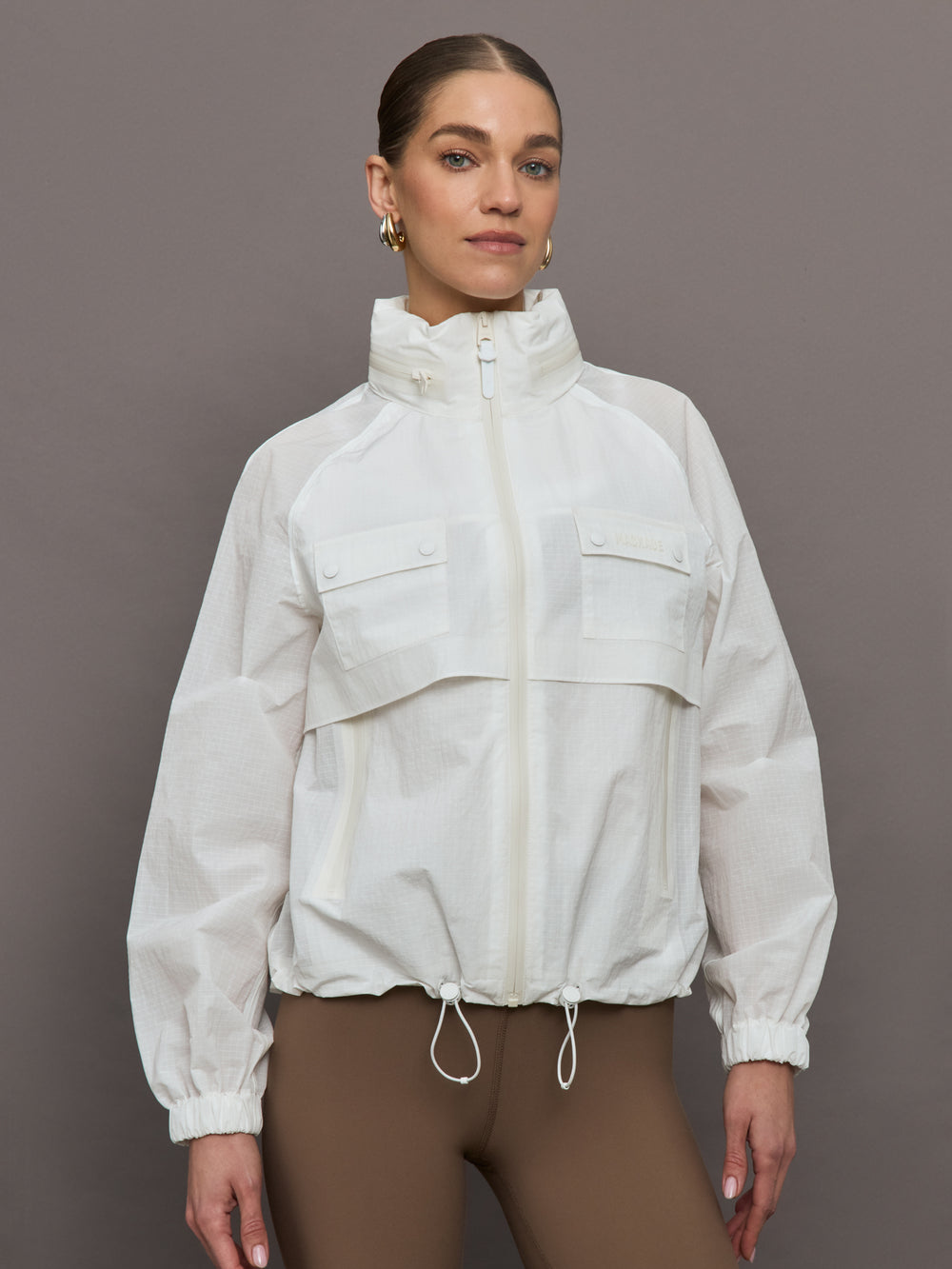 Model is wearing a white windbreaker with drawstrings at the bottom of the jacket and twin chested pockets and a detachable hood. 