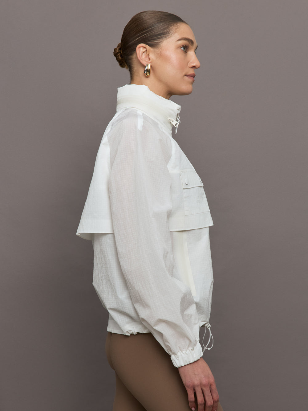 Model is wearing a white windbreaker with drawstrings at the bottom of the jacket and twin chested pockets and a detachable hood. 