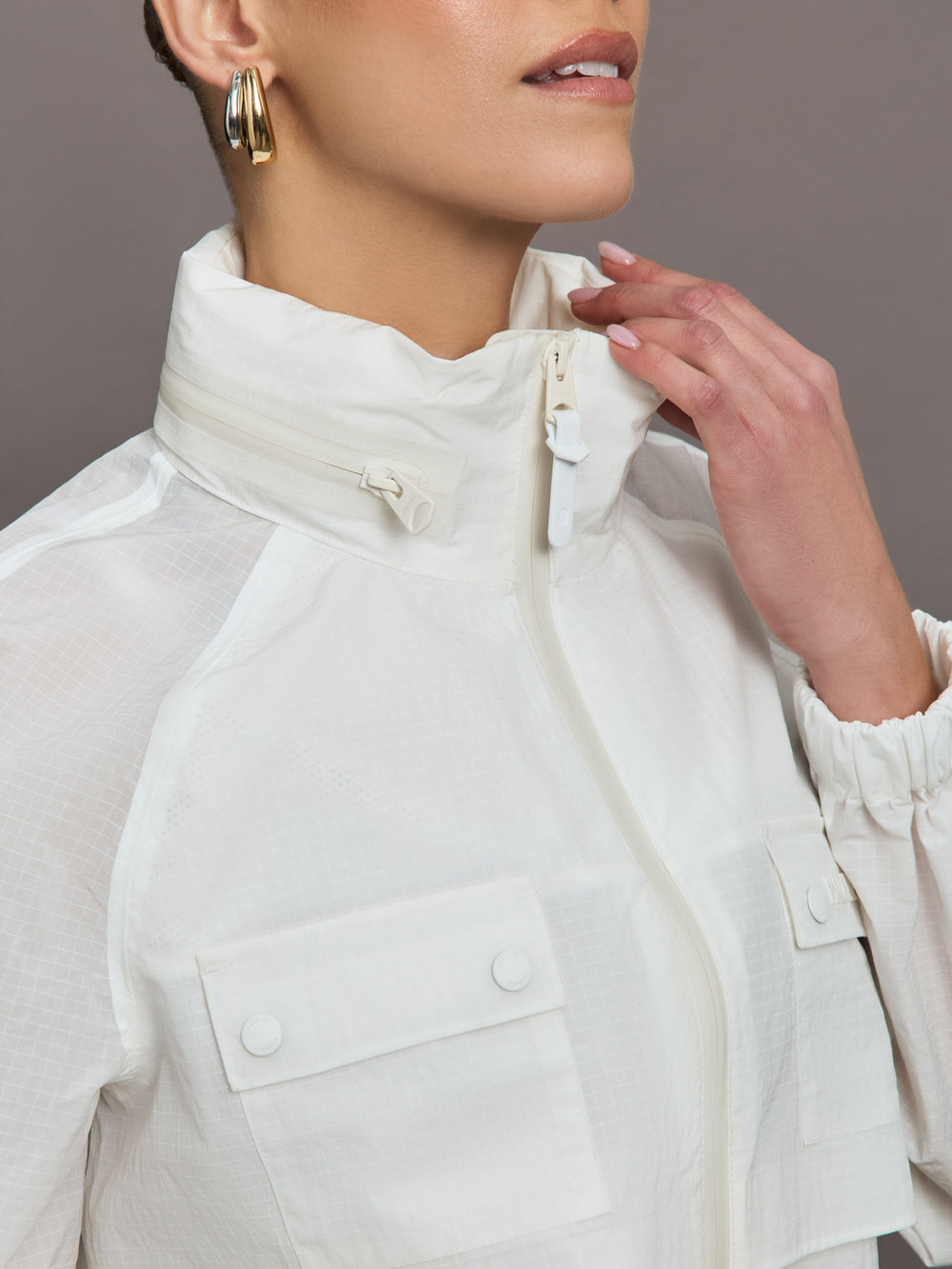 Model is wearing a white windbreaker with drawstrings at the bottom of the jacket and twin chested pockets and a detachable hood. 