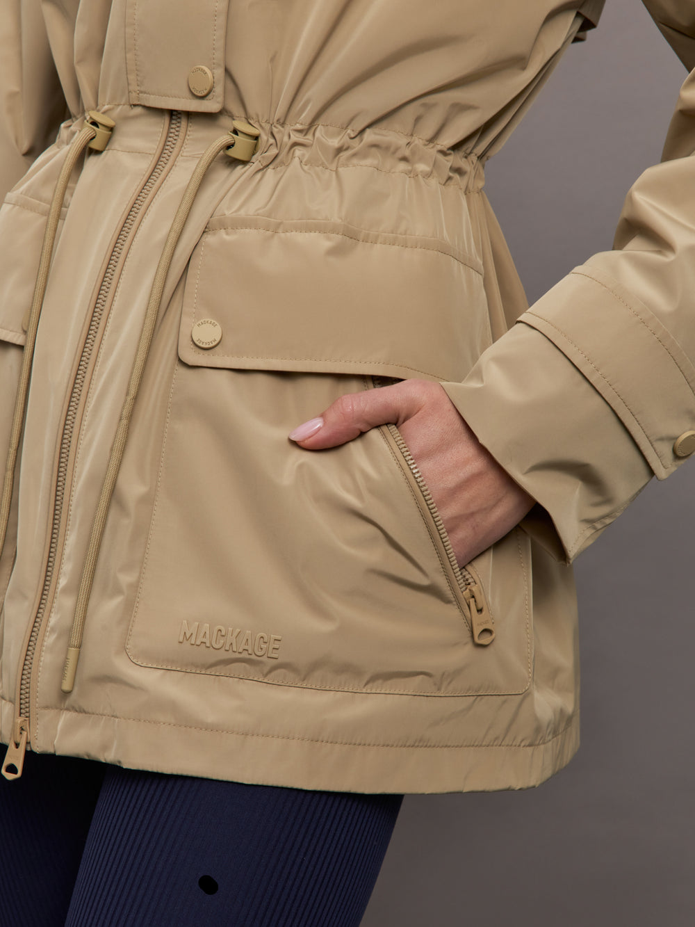 Model is wearing a tan colored rain jacket with a cinched waist.