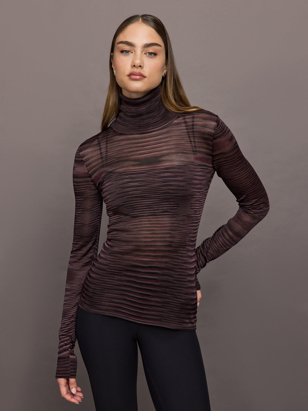 Model wears a striped sheer long sleeve turtleneck with brown and purple tones. 