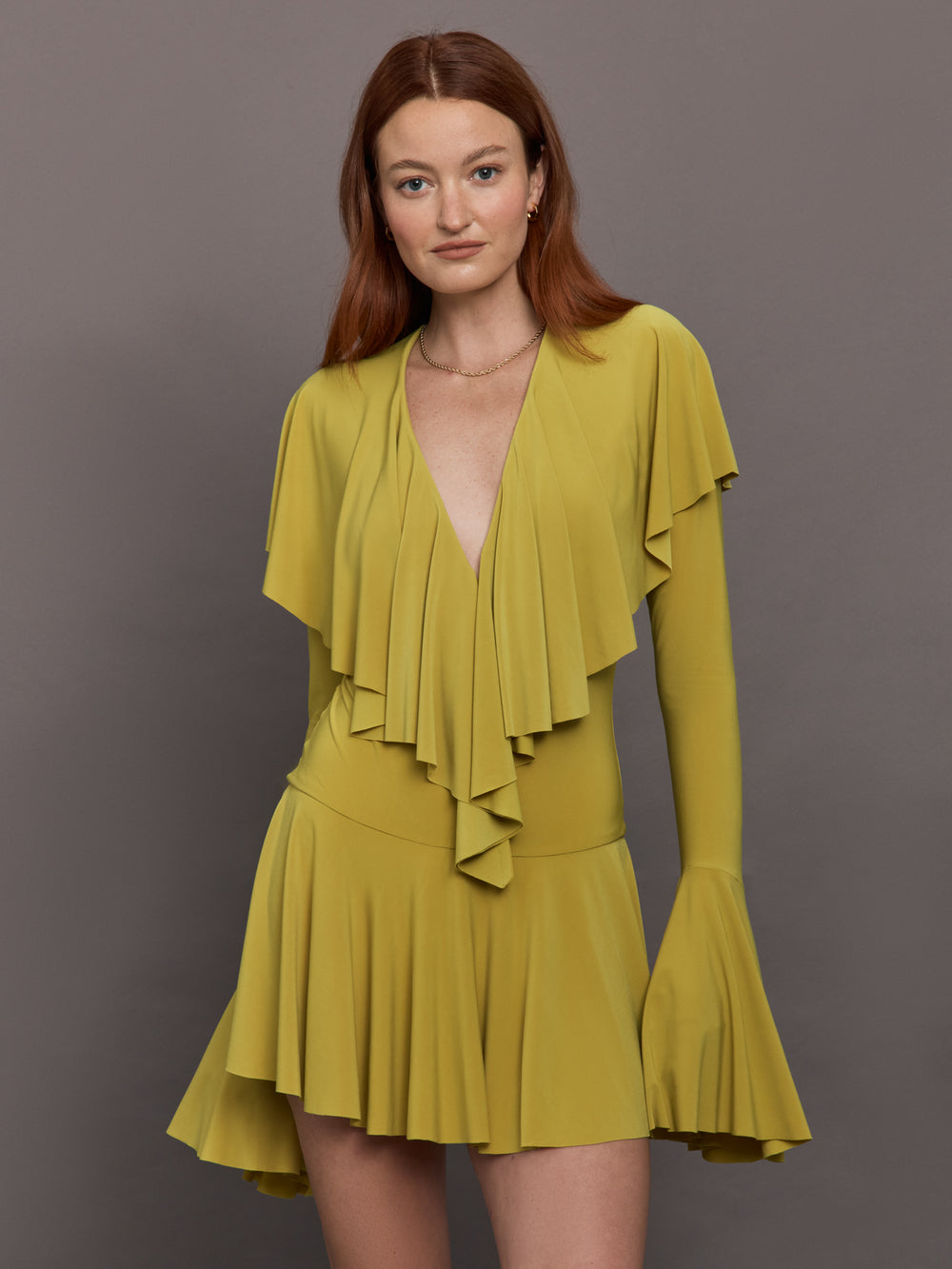 Model wears long sleeve mini dress in asparagus green with a deep V-neckline and ruffle detailing.




