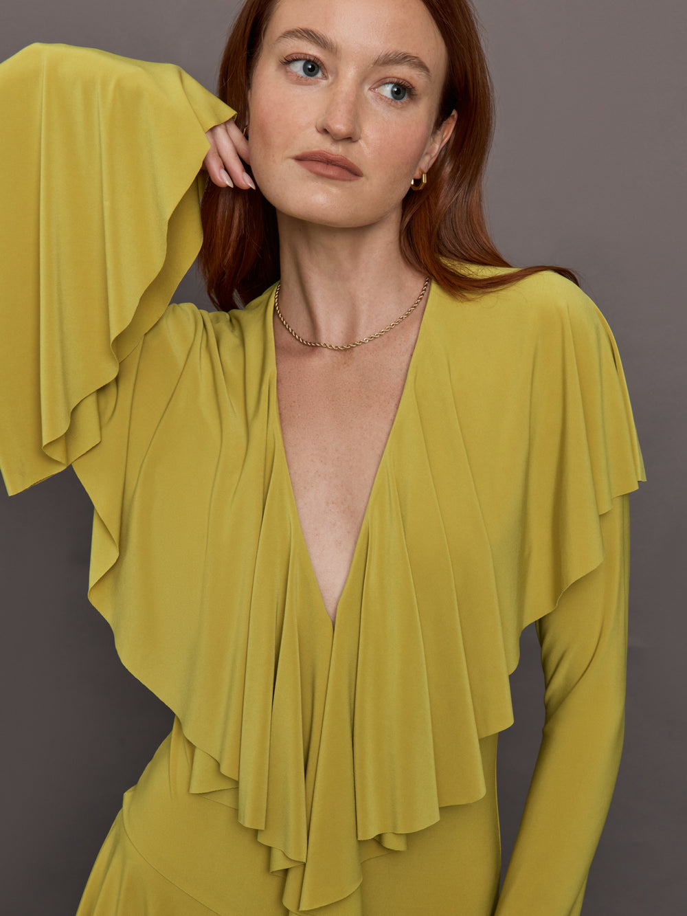 Model wears long sleeve mini dress in asparagus green with a deep V-neckline and ruffle detailing.

