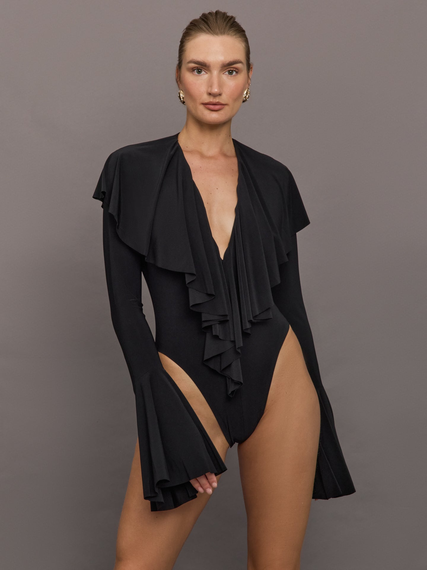 Model wears a black high cut bodysuit with fishtail sleeves, deep V, and a ruffle design.