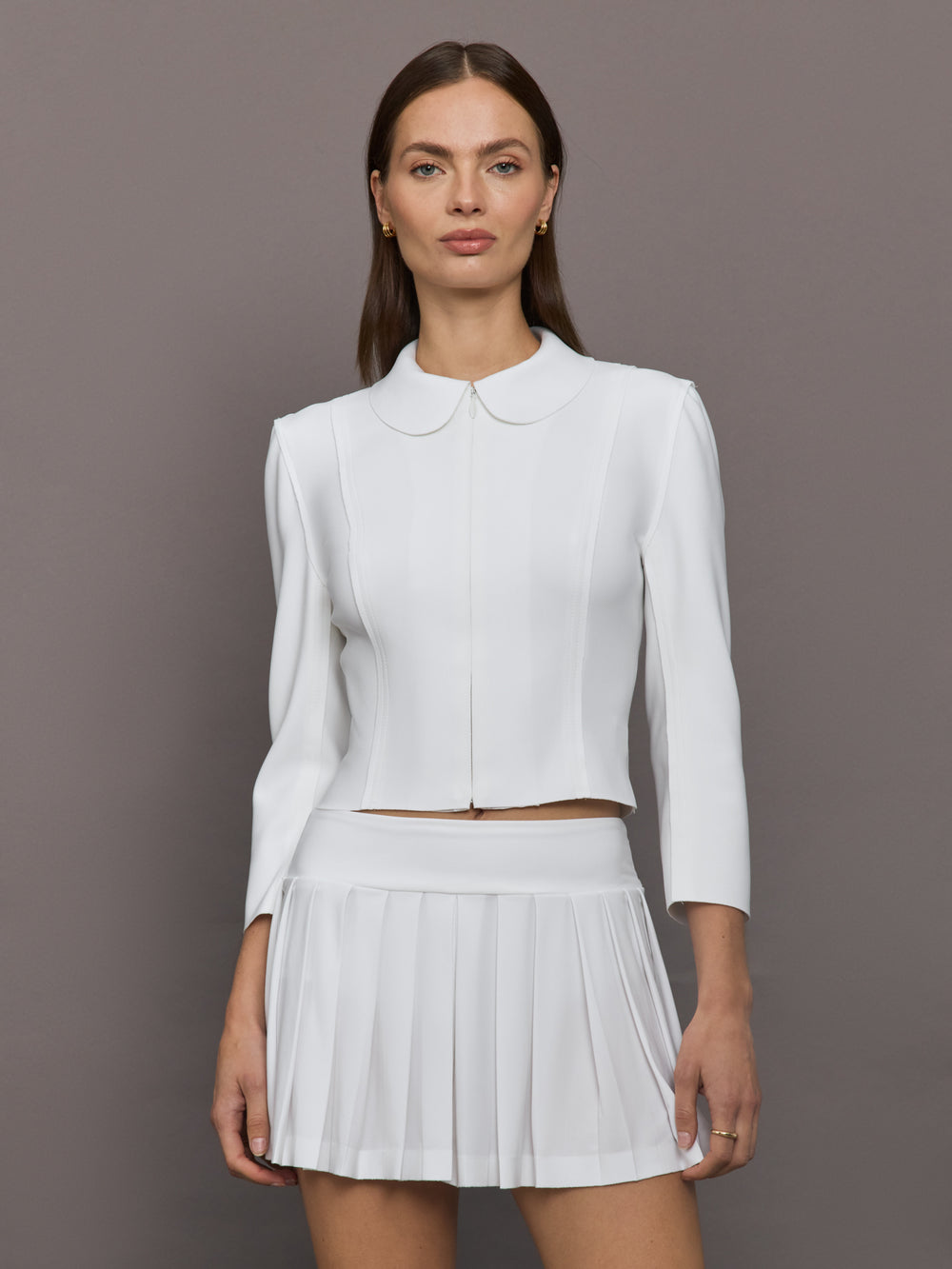 Model wears white jacket with a front zipper, collared neck and long sleeves.




