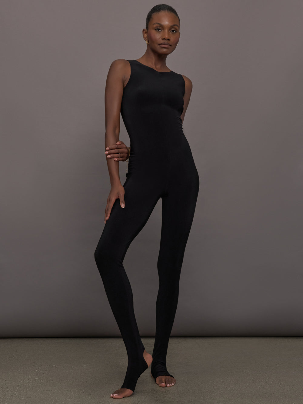 Model wears a black sleeveless X catsuit with built-in footies, featuring a high neckline, body-contouring fit, and crisscross X straps on the back