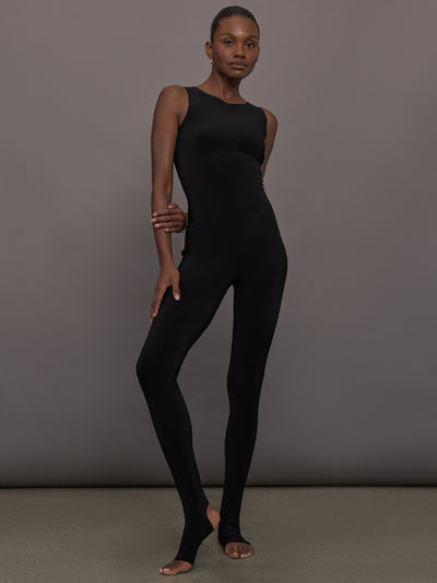 Model wears a black sleeveless X catsuit with built-in footies, featuring a high neckline, body-contouring fit, and crisscross X straps on the back