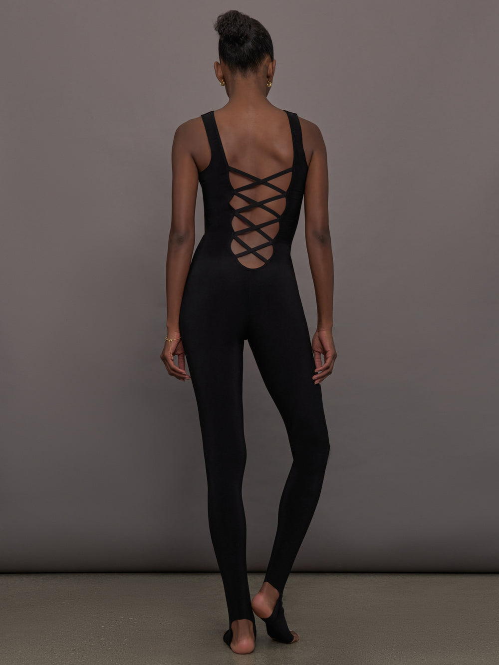 Model wears a black sleeveless X catsuit with built-in footies, featuring a high neckline, body-contouring fit, and crisscross X straps on the back