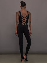 Model wears a black sleeveless X catsuit with built-in footies, featuring a high neckline, body-contouring fit, and crisscross X straps on the back