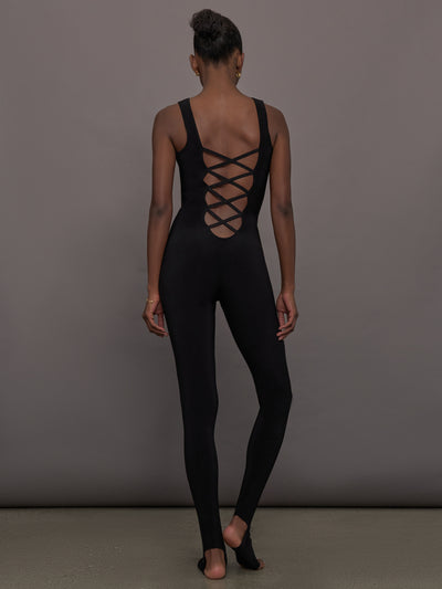 Model wears a black sleeveless X catsuit with built-in footies, featuring a high neckline, body-contouring fit, and crisscross X straps on the back