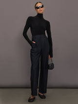 Person wearing a black turtleneck and high-waisted pants against a gray background