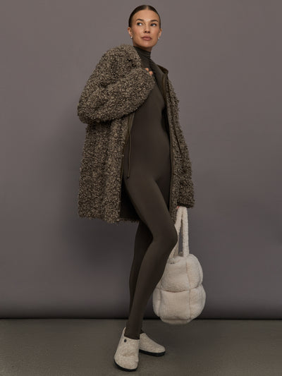 Woman wearing a brown textured coat, brown outfit, and white shoes holding a white bag against a gray background