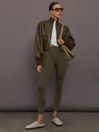 Woman wearing a brown jacket, white shirt, green leggings, and gray slippers against a gray background.