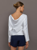 Woman wearing a white boatneck long sleeve blouse and navy shorts against a gray background