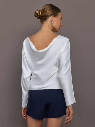 Woman wearing a white boatneck long sleeve blouse and navy shorts against a gray background