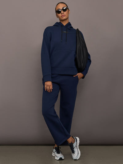Model is wearing On Focus Tech Hoodie for women in navy styled with matching joggers, sneakers, sunglasses, and black shoulder bag, full front view.