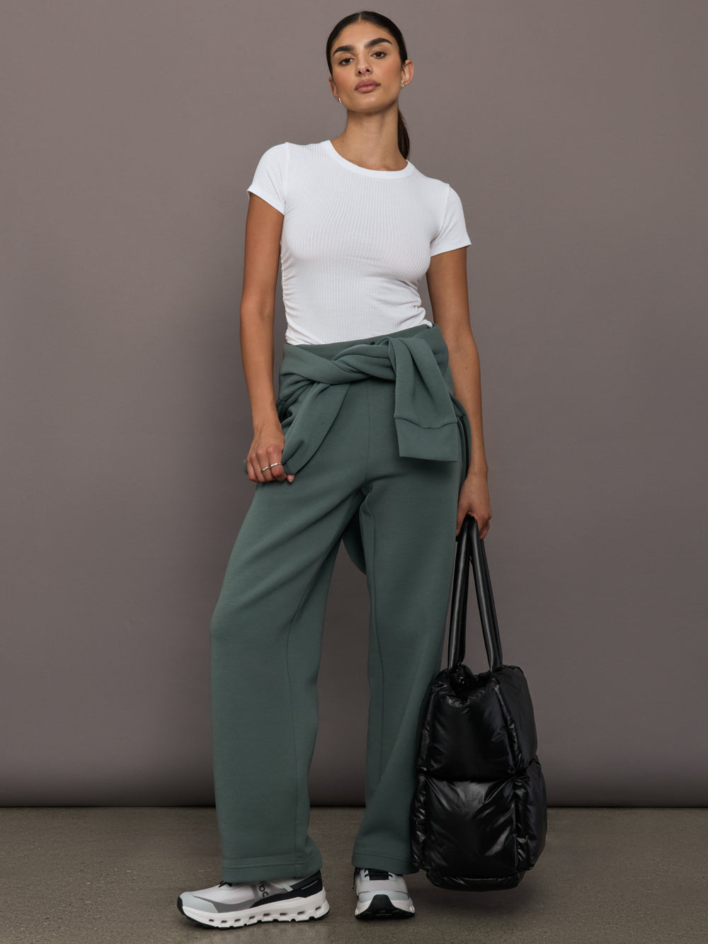 Woman wearing a white t-shirt and green pants holding a black bag against a gray background