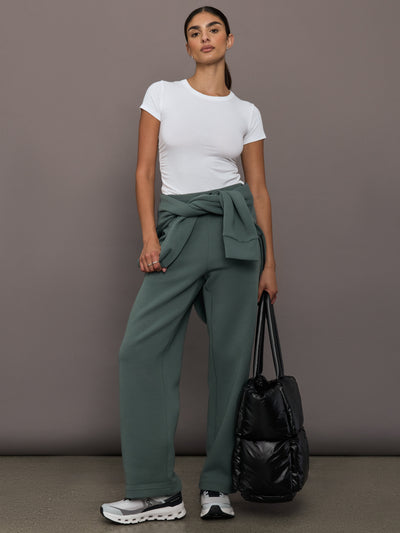 Woman wearing a white t-shirt and green pants holding a black bag against a gray background