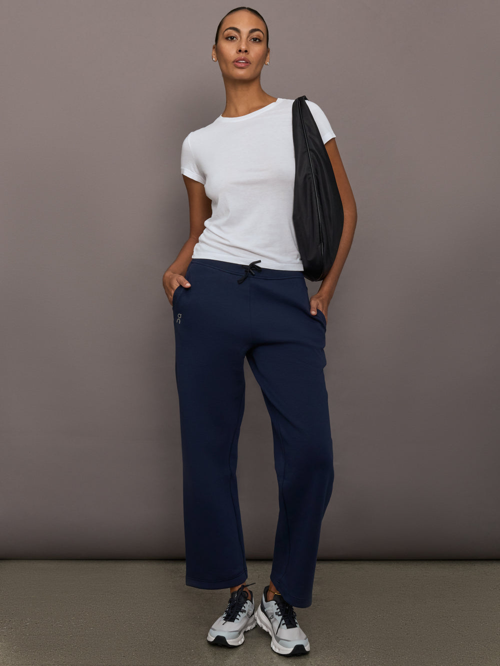 Model is wearing On Focus Tech Sweatpants for women in navy styled with white t-shirt, sneakers, and black shoulder bag, full front view.