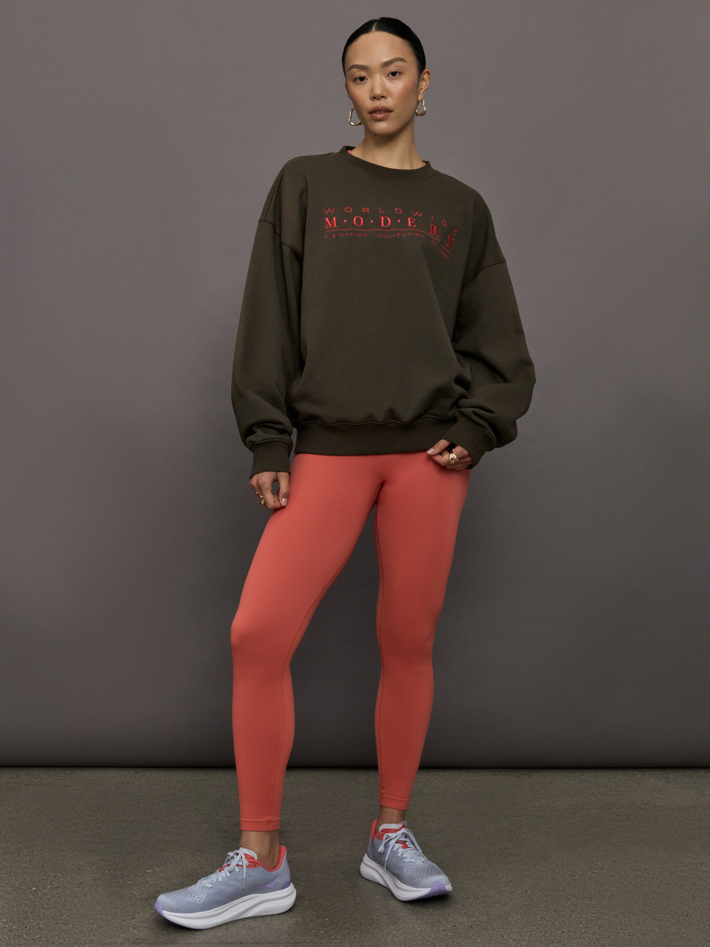 Model wears an oversized brown crewneck sweatshirt with the words 'Worldwide Modern' printed in coral coloring across the chest.