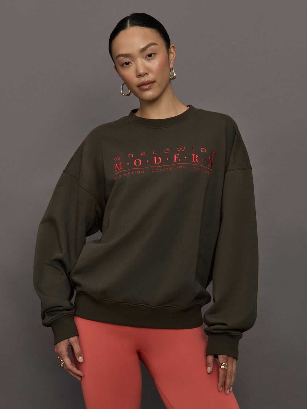 Model wears an oversized brown crewneck sweatshirt with the words 'Worldwide Modern' printed in coral coloring across the chest.